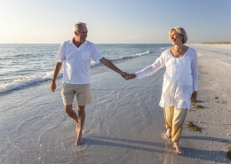 Happy senior man and woman couple walking and holding hands on a deserted tropical beach with bright clear blue sky after retiring abroad