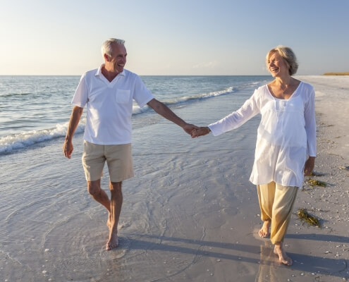 Happy senior man and woman couple walking and holding hands on a deserted tropical beach with bright clear blue sky after retiring abroad