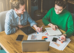 Teamwork. Two smiling young business women sitting in office at table and working. On table is laptop, smartphone, tablet, paper charts and notebook. Students learning online, chatting. Brainstorming.