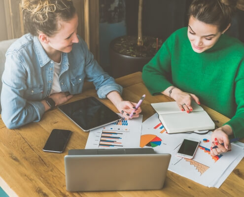 Teamwork. Two smiling young business women sitting in office at table and working. On table is laptop, smartphone, tablet, paper charts and notebook. Students learning online, chatting. Brainstorming.