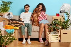 taking pet abroad concept. Happy diverse couple sit on sofa with favourite dog, have break on moving day, busy unpacking, have mess in new flat, many packages, remove to own house