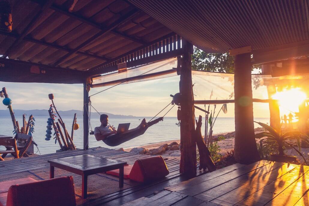 Young attractive guy a man with a beard in a hammock with a laptop on the background of the sea and sunset, distant work, freelancer, blogger, vacation and travel